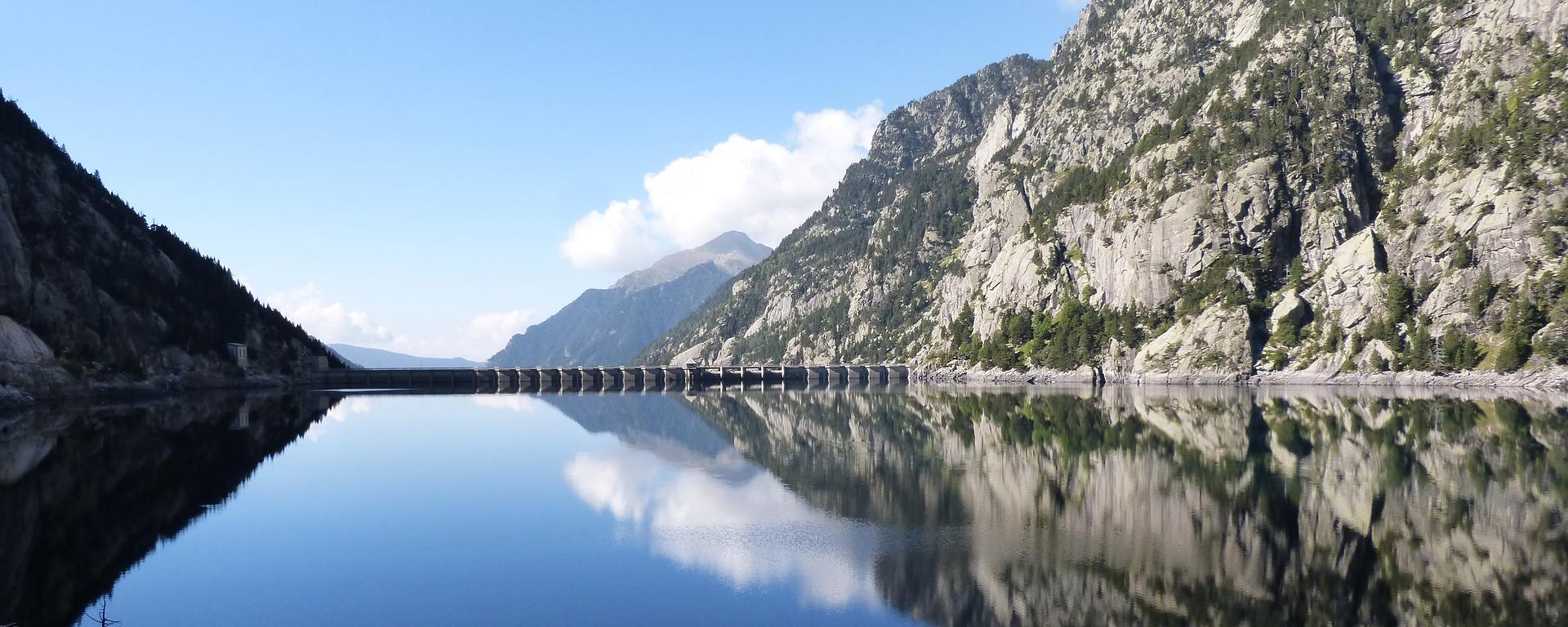 A calm blue lake nestled between three mountains. There is a bridge/hydroelectric dam in the distance